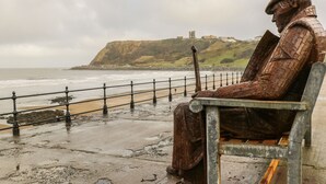 Una statua raffigura una figura seduta con una spada, che domina una spiaggia con un promontorio roccioso e un castello in lontananza.