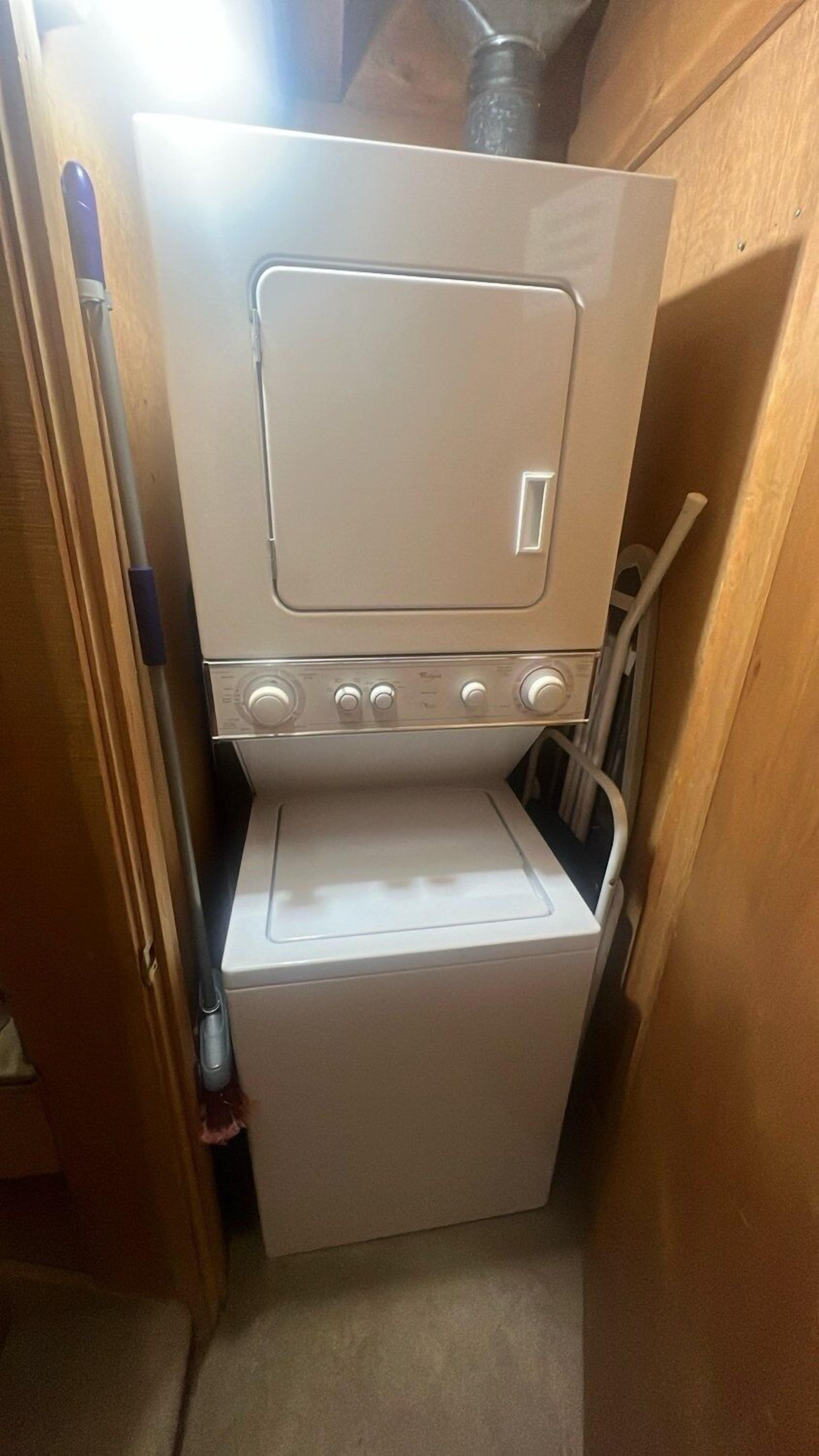 A compact, white laundry machine in a small, enclosed space with a wooden door and a mop leaning against the wall.