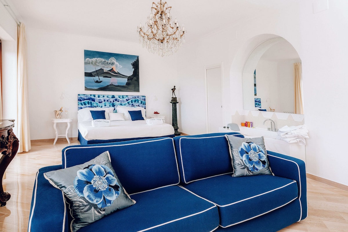 A modern hotel room with a blue sofa, a bed with white bedding, a painting of a coastal scene, a chandelier, and a bathroom with a mirror and sink.