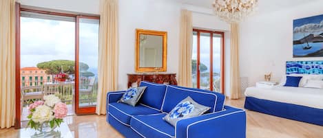 A modern hotel room with a blue sofa, a glass coffee table, a bed, and a large window with a view of the city.