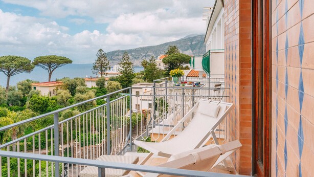 Un balcon avec des fauteuils de jardin blancs, une vue sur la mer et, au loin, des maisons à étages.