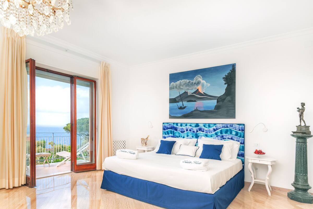 A bedroom with a bed, a painting of a volcano, a statue, and a view of the sea.