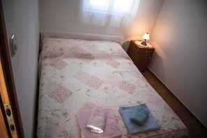 Una cama pequeña y bien hecha con una colcha estampada con motivos florales, una mesita de noche con una lámpara y dos toallas dobladas sobre la cama.
