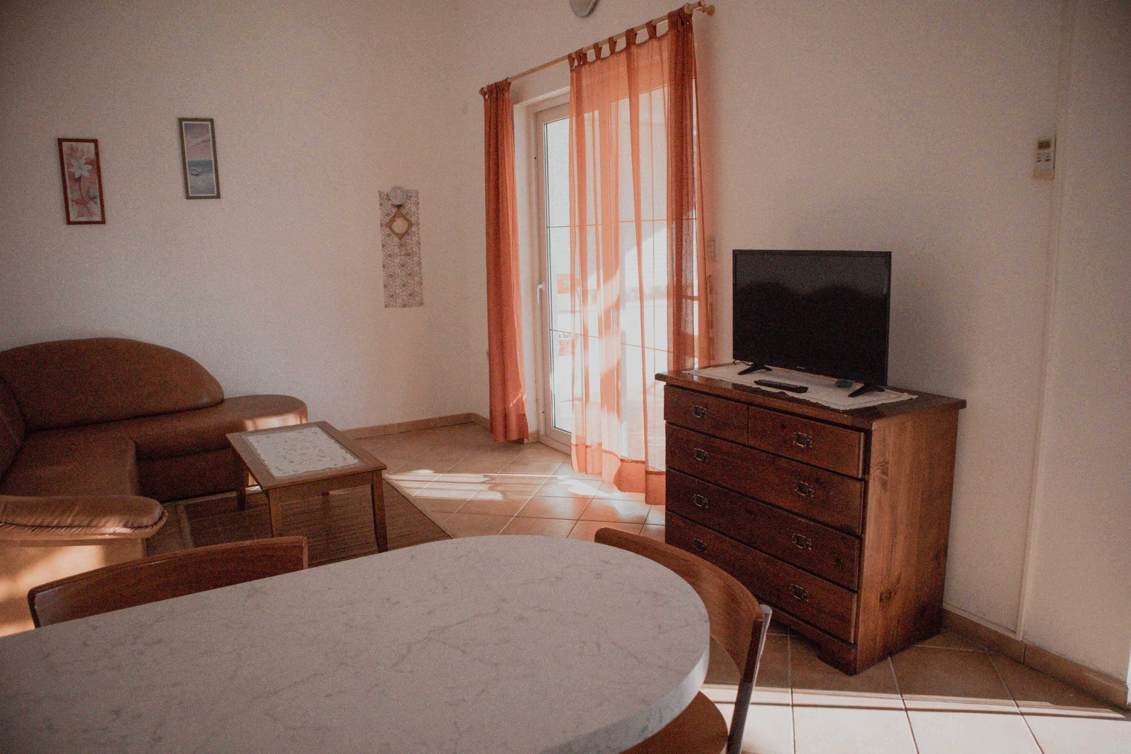 A living room with a sofa, a TV on a wooden cabinet, a table, and a window with curtains.