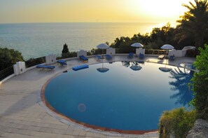 A circular infinity pool with lounge chairs and umbrellas, overlooking a body of water at sunset.