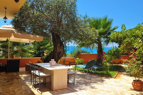 An outdoor patio with a table and chairs, surrounded by lush greenery and a view of the sea. - House Angelos D With sea View and Private Garden - Agios Gordios Beach (Corfu)