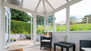 Une véranda dotée d’une porte vitrée donnant sur un patio aménagé avec du mobilier d’extérieur et offrant une vue sur un jardin verdoyant.