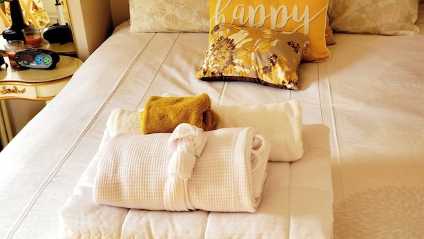 Cama arrumada com um travesseiro que diz 'happy', uma toalha dobrada e um relógio em uma mesa de cabeceira.