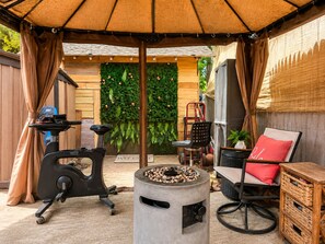 An outdoor patio area with a stationary exercise bike, a fire pit, a seating area with a red cushion, and a wall with green foliage. - Room-yellow Room Near Yale (New Haven)