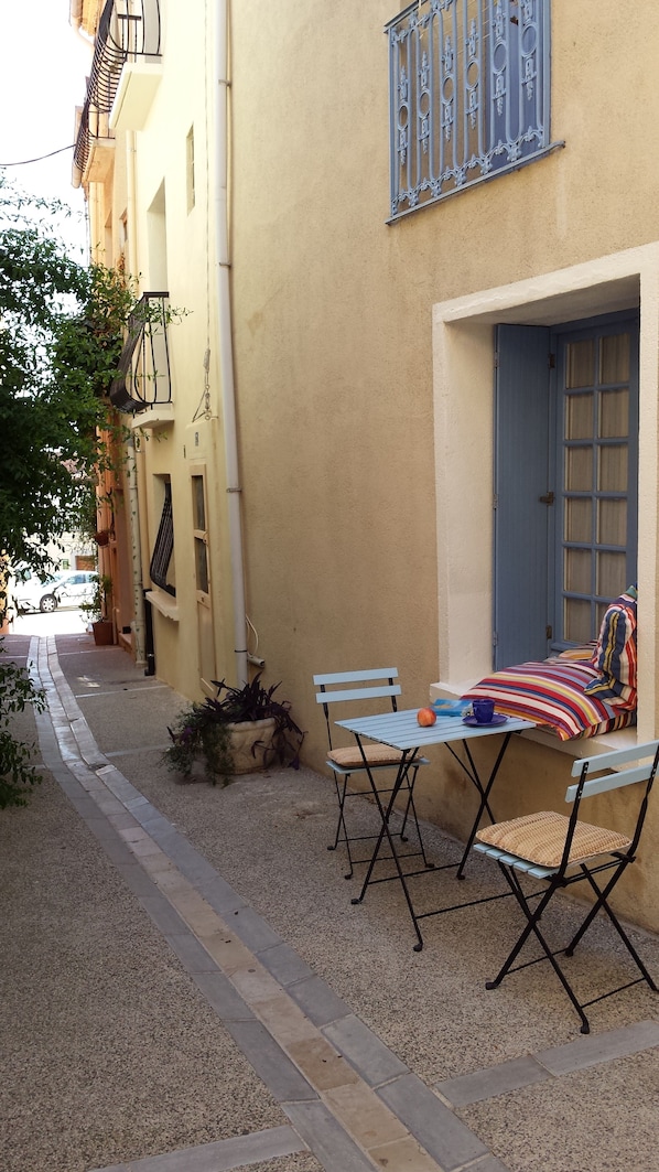 A narrow street with a building featuring a blue door and window, a small table with a striped cushion, and a bench with a woven seat.
