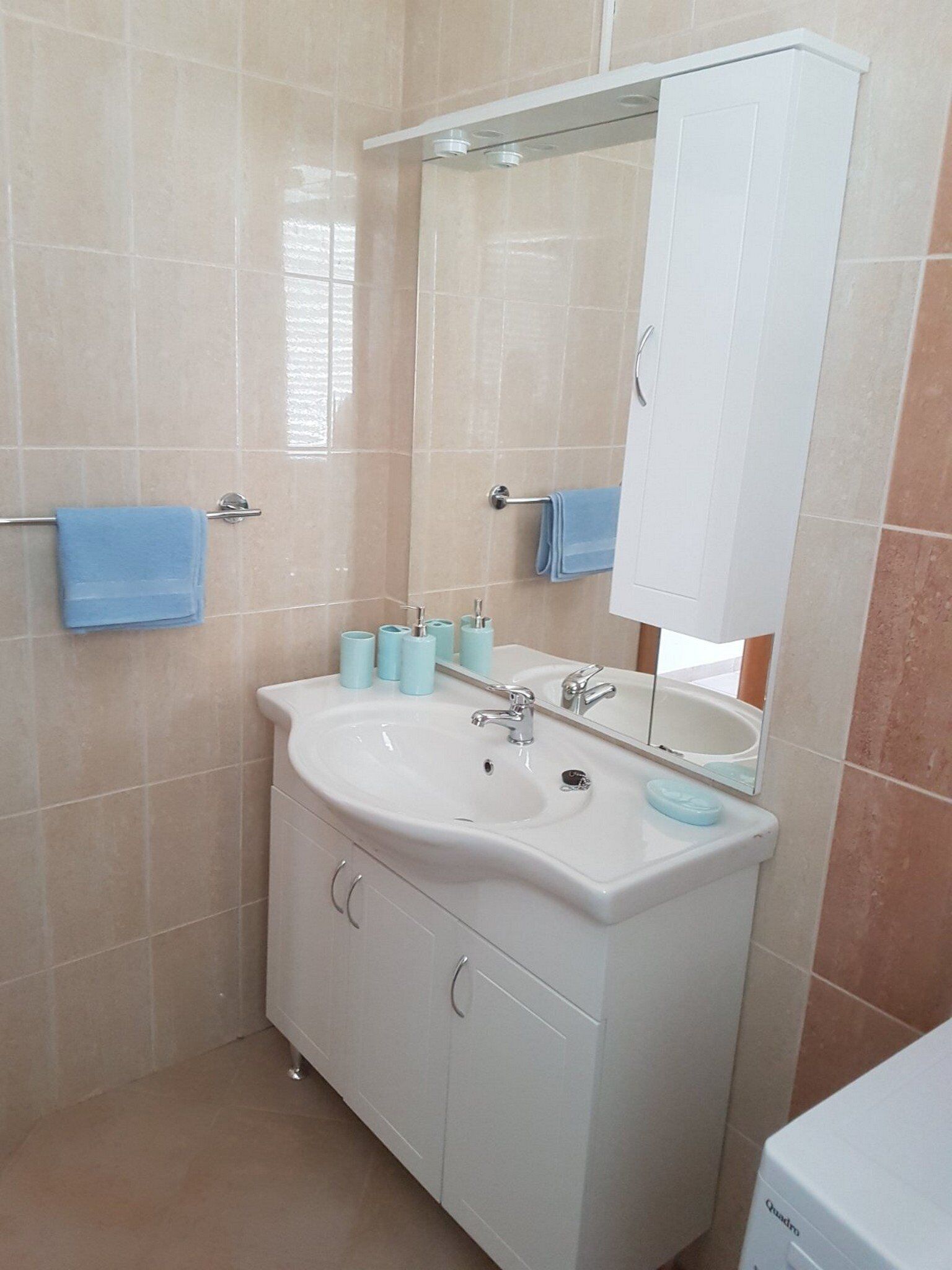 Een badkamer met een wit wastafelmeubel, spiegel en kastje, beige tegels en twee blauwe handdoeken.