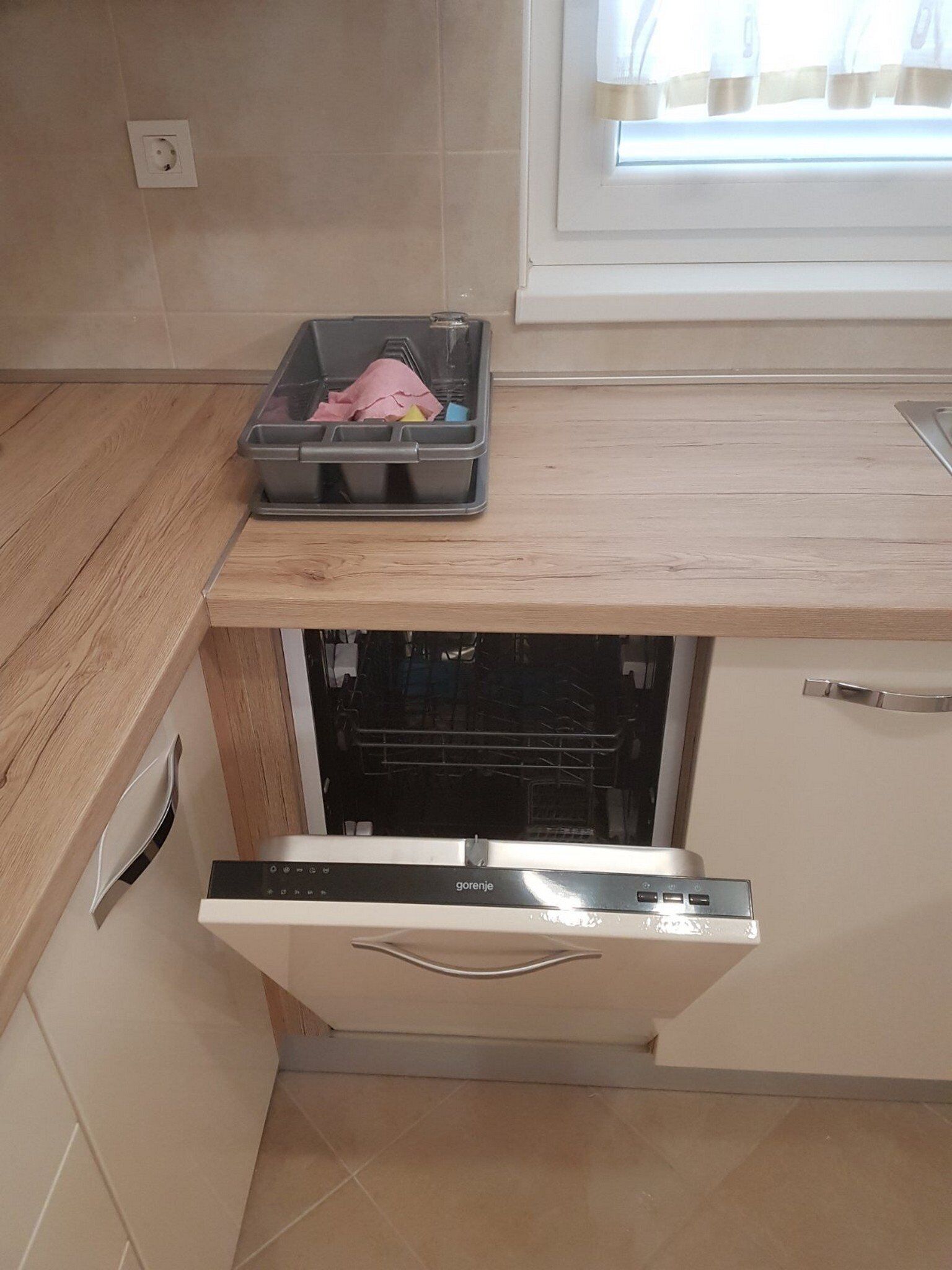 A compact kitchen with a built-in dishwasher, a window with blinds, and a small countertop with a plastic container.
