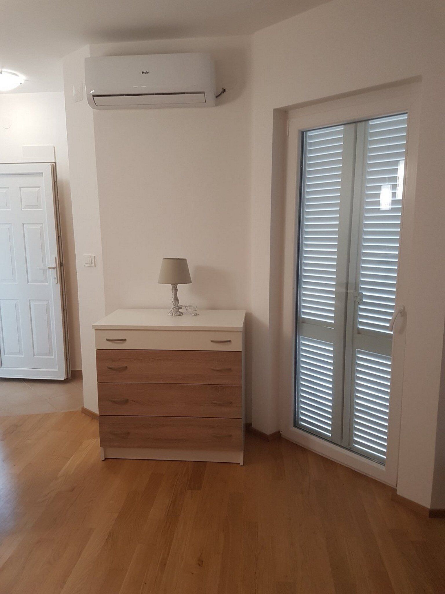 Ein Zimmer mit einer weißen Klimaanlage, einem Holzschrank mit Schubladen, einer Lampe und einer Glastür mit Jalousien.