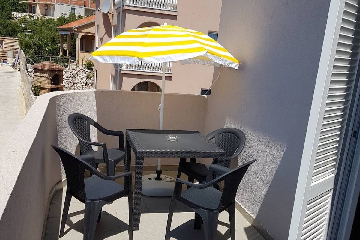 An outdoor patio with a table and chairs, a yellow and white striped umbrella, and a building with a balcony in the background.