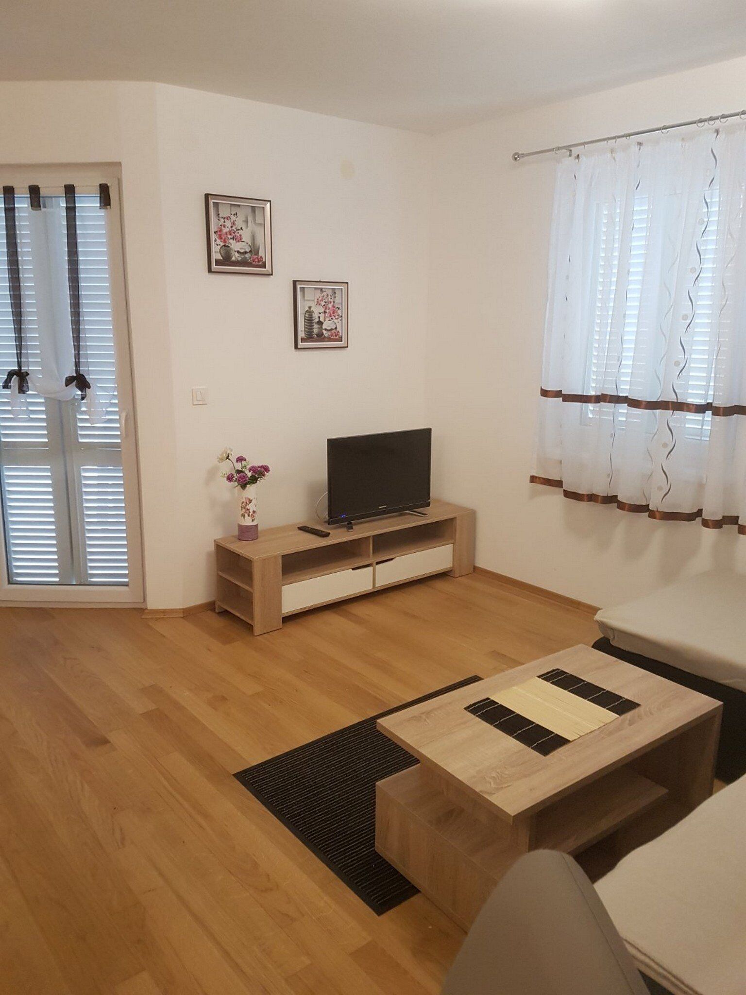 A living room with a wooden floor, a TV on a wooden stand, a sofa, a coffee table, and a window with curtains.