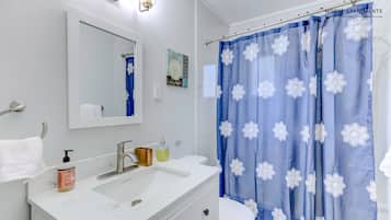 Un baño con lavamanos blanco, espejo, inodoro y cortina de ducha azul con motivos florales blancos.