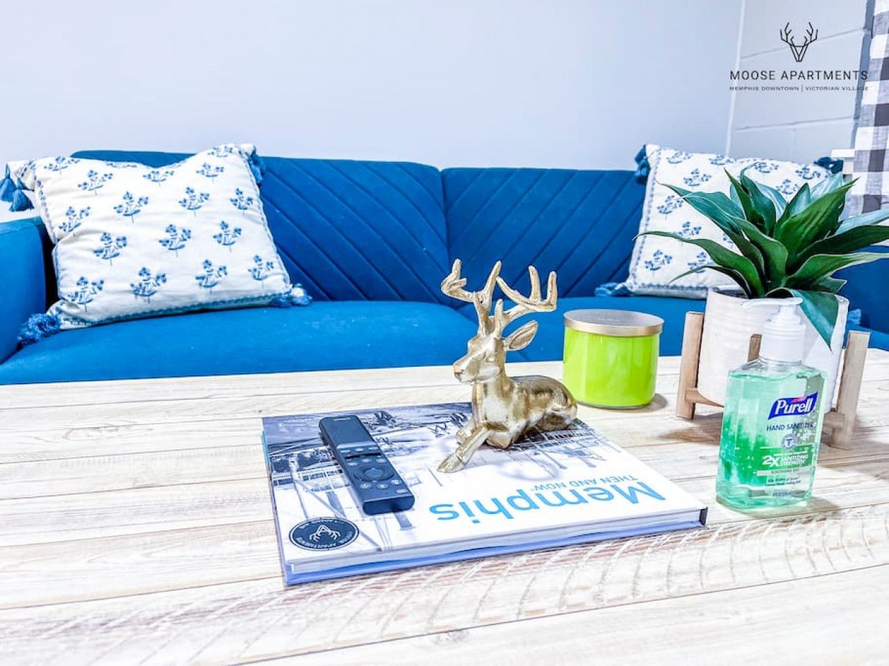 Un canapé bleu orné de coussins à motifs, une petite figurine de cerf, une télécommande, un magazine, une bougie verte et un flacon de gel hydroalcoolique, le tout posé sur une table en bois.