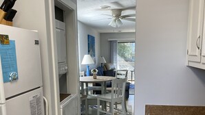 Een compacte keuken met witte kastjes, een inbouwoven en een koelkast. Er is een eethoek met een tafel en stoelen, een plafondventilator en uitzicht door een glazen deur.