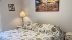 Een slaapkamer met een bed, een plafondventilator, een lamp, een nachtkastje en een schilderij van een strand.