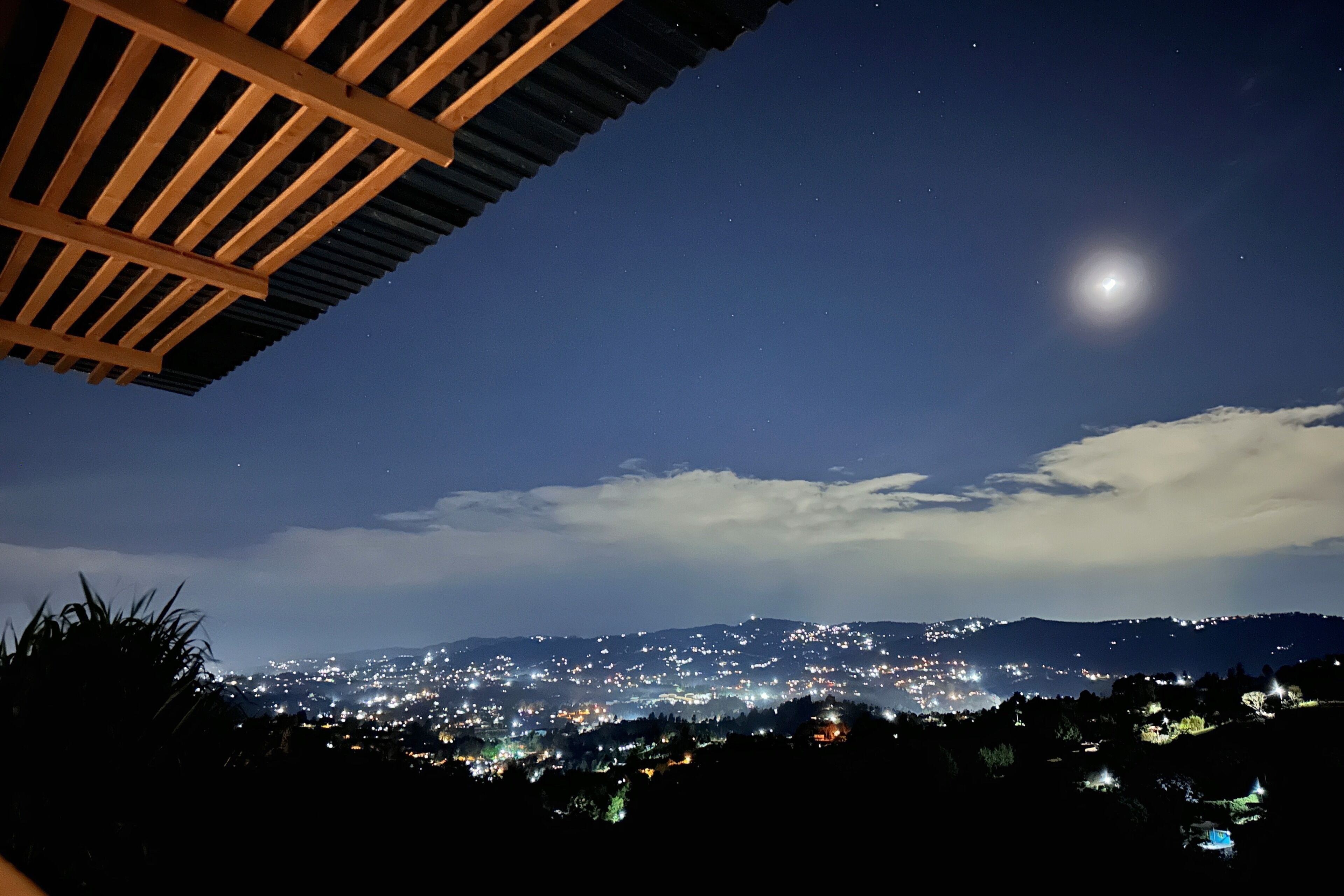 Vista nocturna de una ciudad con una luna destacada en el cielo, una estructura de madera en primer plano y un horizonte lejano iluminado.