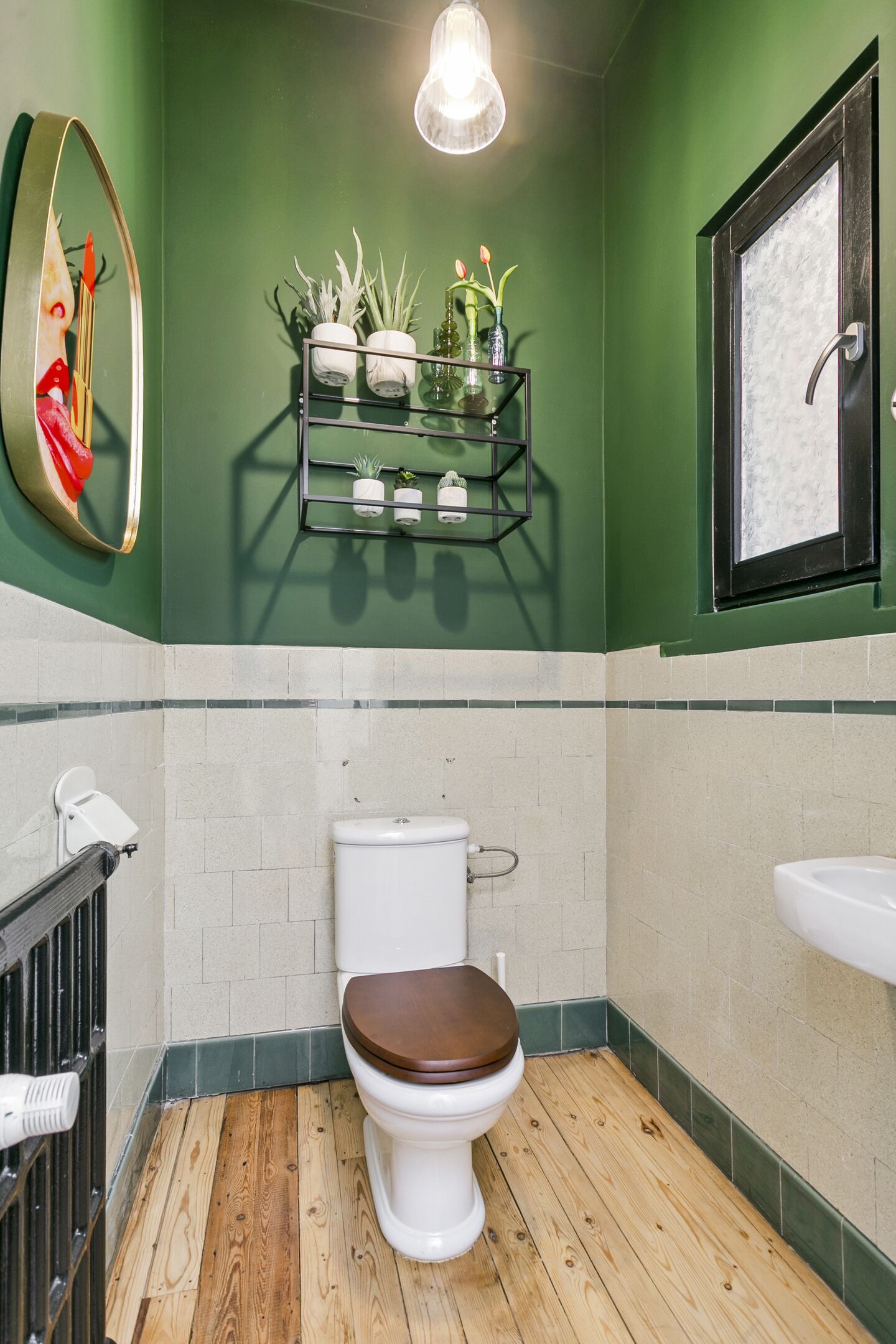 A bathroom with a wooden floor, a white toilet, a shelf with plants, and a mirror with a red object.