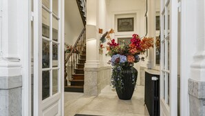Un pasillo con suelo de mármol, un gran jarro negro con flores, una escalera y un radiador negro.