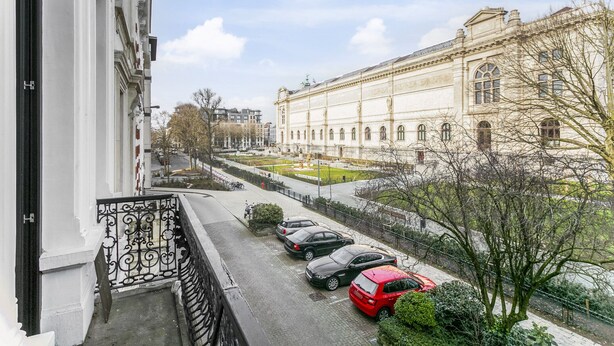 Vista de la calle con coches aparcados, un edificio de arquitectura clásica y cielo despejado.