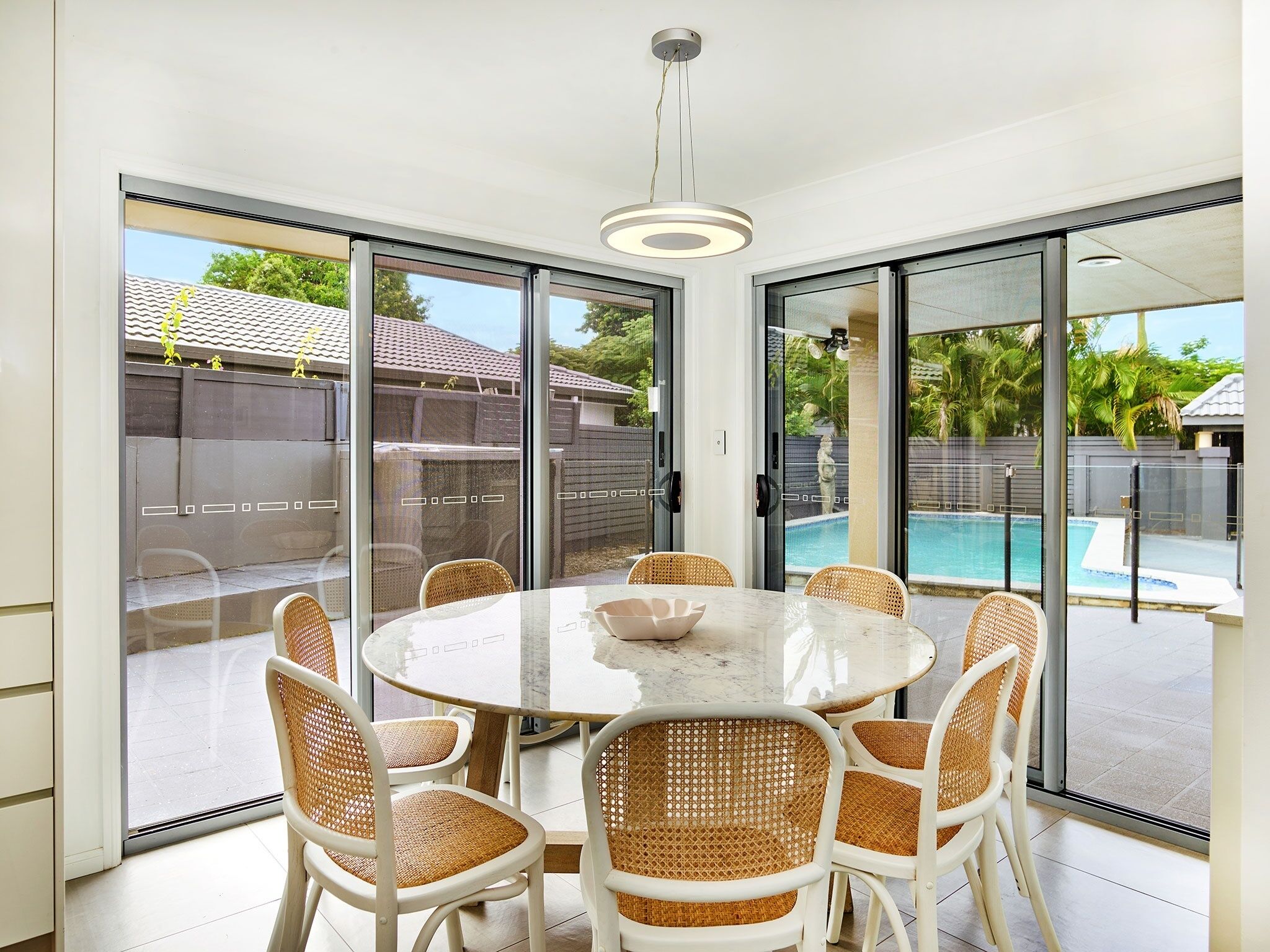 A modern dining area with a round table and wicker chairs, large sliding glass doors leading to a pool.