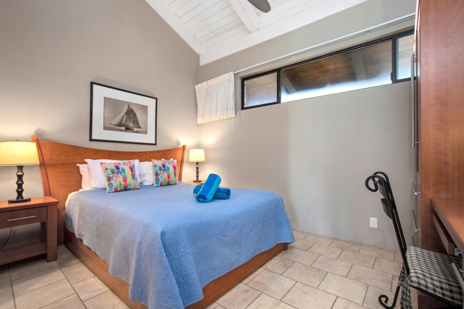 Une chambre à coucher comprenant un lit, deux tables de chevet, une chaise, une fenêtre avec des stores et un ventilateur de plafond.