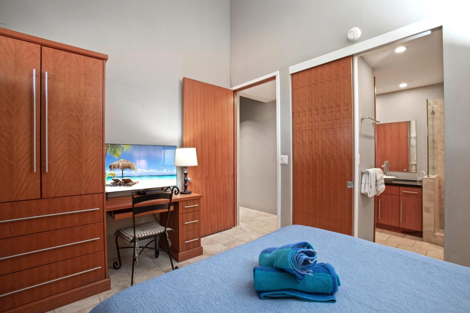 Ein Hotelzimmer mit einem Bett, einem Schreibtisch mit Computer, einem Fernseher, der eine Strandansicht zeigt, einem Badezimmer mit Spiegel und Waschbecken sowie Holzschränken.