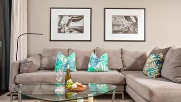 Un salon avec un canapé beige, deux tableaux encadrés aux motifs nautiques en noir et blanc, une table basse en verre sur laquelle se trouvent une bouteille de vin, des raisins et du fromage, ainsi que des coussins aux motifs tropicaux.