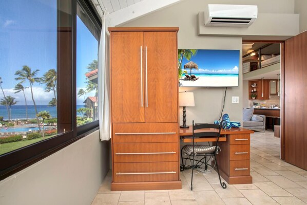 Une pièce avec une armoire en bois, un bureau avec une chaise, un téléviseur fixé au mur et une vue sur la plage par la fenêtre.