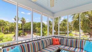 A covered outdoor seating area with a striped sofa and a small table, overlooking a lush garden with palm trees. - Pelican Bay 13 - Brand New Reno! 1 Block to Beach! (Isle of Palms)