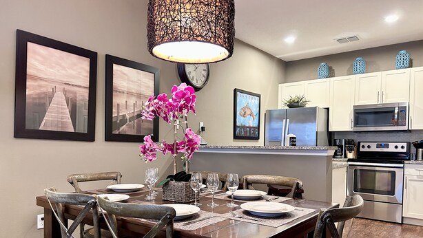 Un comedor con una mesa de madera, sillas y un jarrón de flores rosas. Se observan cuadros en la pared, un reloj y armarios de cocina al fondo.