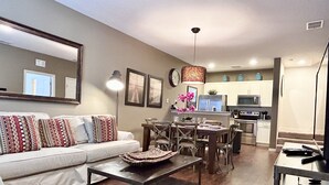 Una sala de estar con sofá, mesa de centro y zona de comedor con sillas y mesa. Se ve una cocina al fondo con armarios y electrodomésticos.