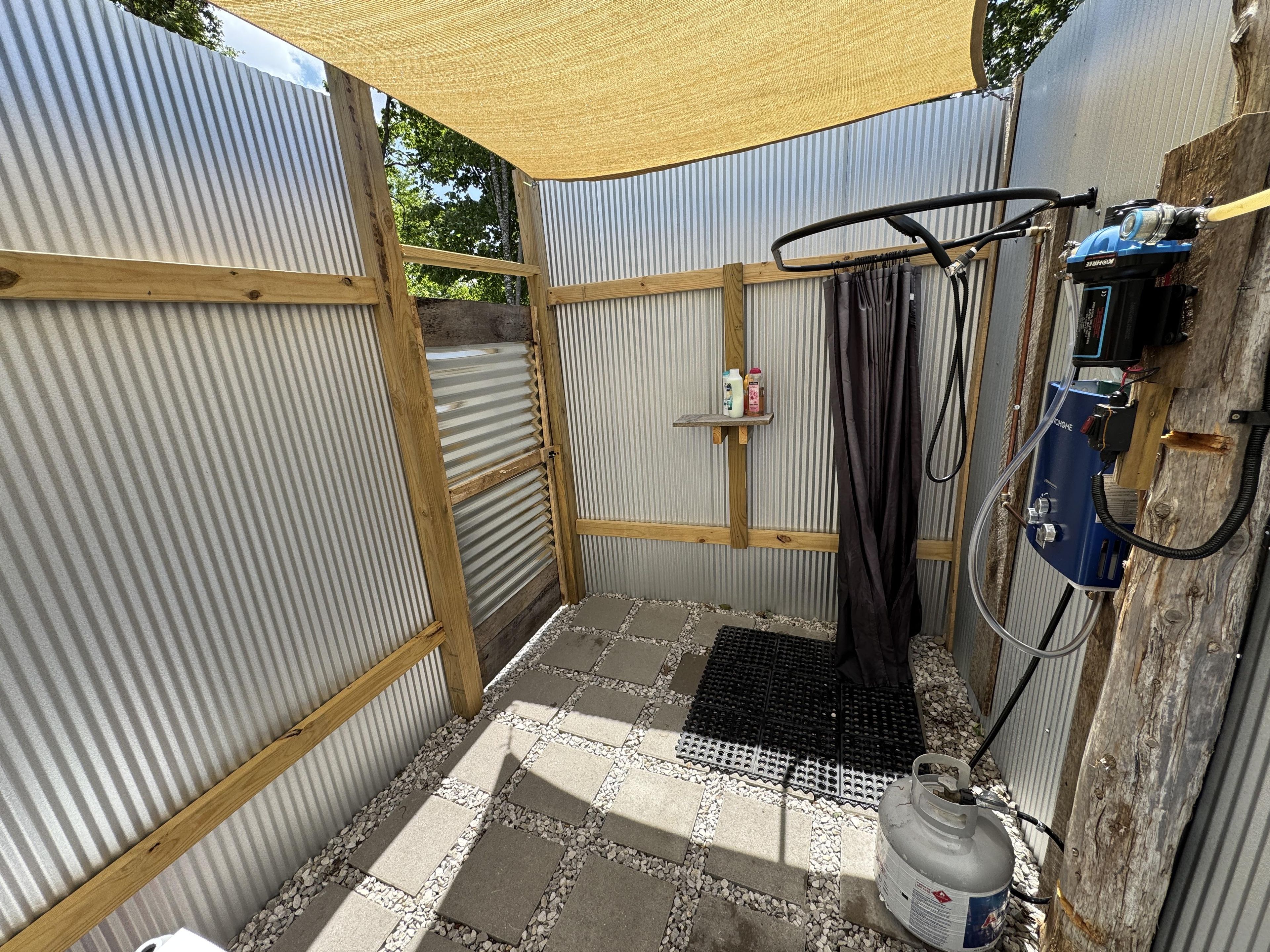 A small, enclosed outdoor shower area with a corrugated metal wall, a wooden frame, a black mat, and a gas cylinder.