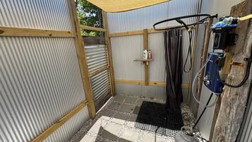 A small, enclosed outdoor shower area with a corrugated metal wall, a wooden frame, a black mat, and a gas cylinder.