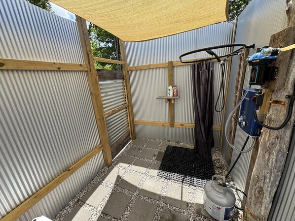 A small, enclosed outdoor shower area with a corrugated metal wall, a wooden frame, a black mat, and a gas cylinder.