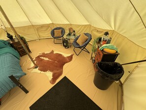 A tent interior with a bed, two chairs, a rug, a waste bin, and a small table with items.