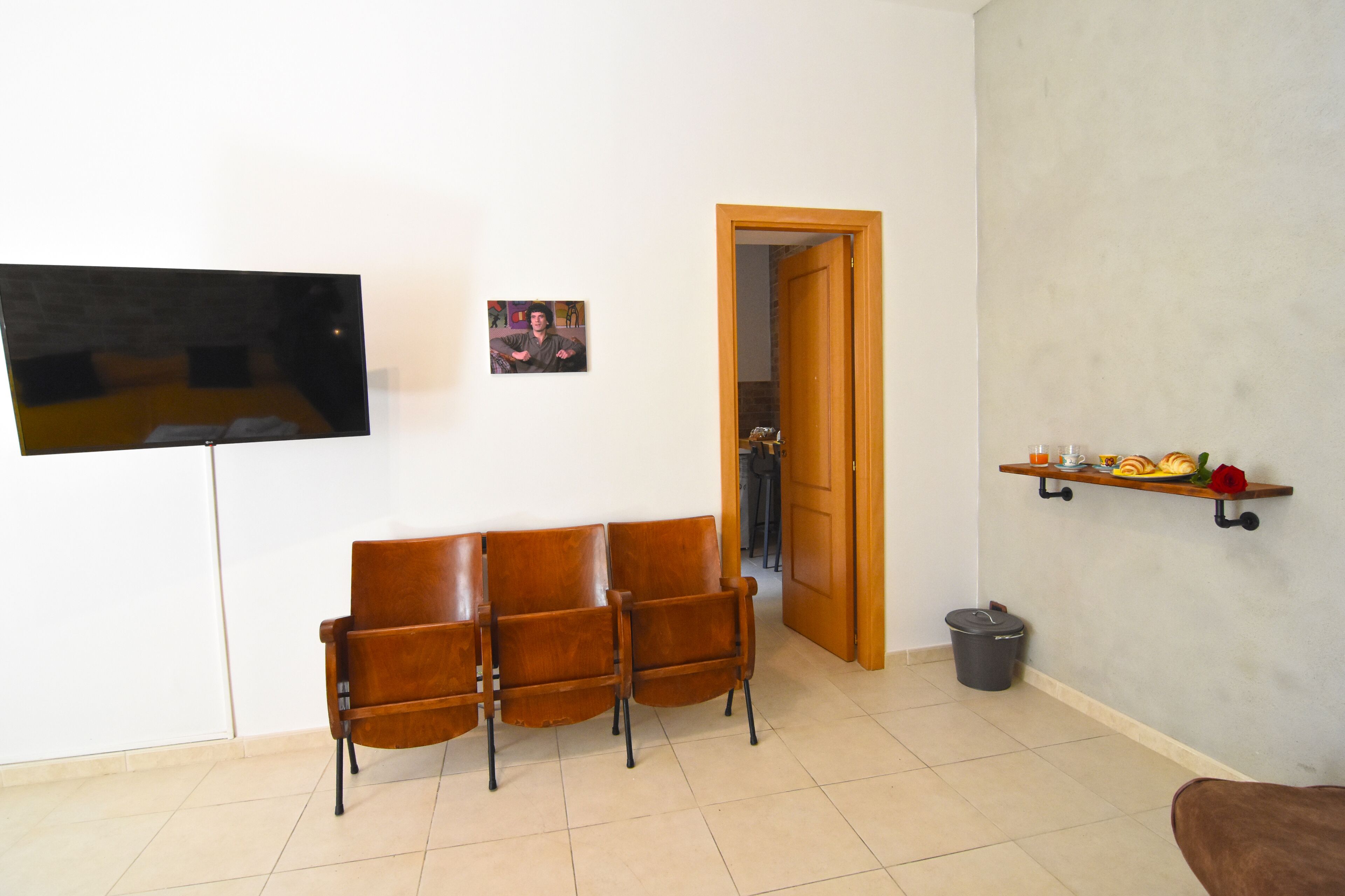 A living room with a TV, three chairs, a wooden door, a trash bin, and a shelf with food items.