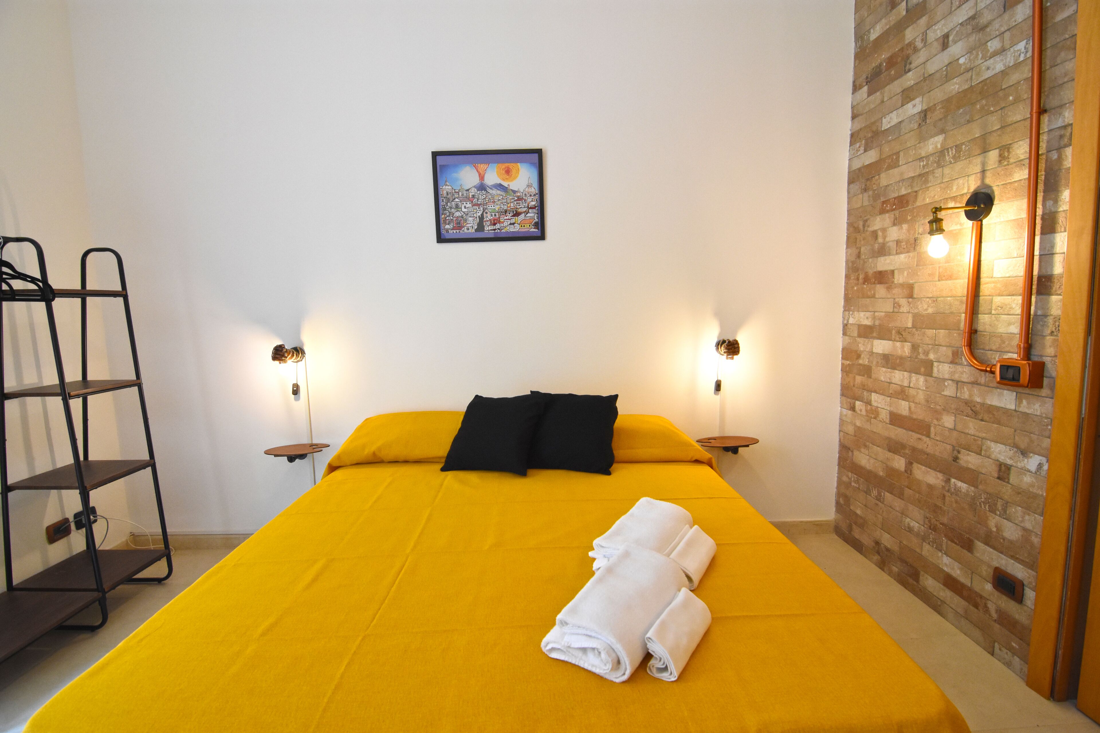 A hotel room with a yellow bed, black pillows, a folded towel, a metal shelf, and a brick wall.