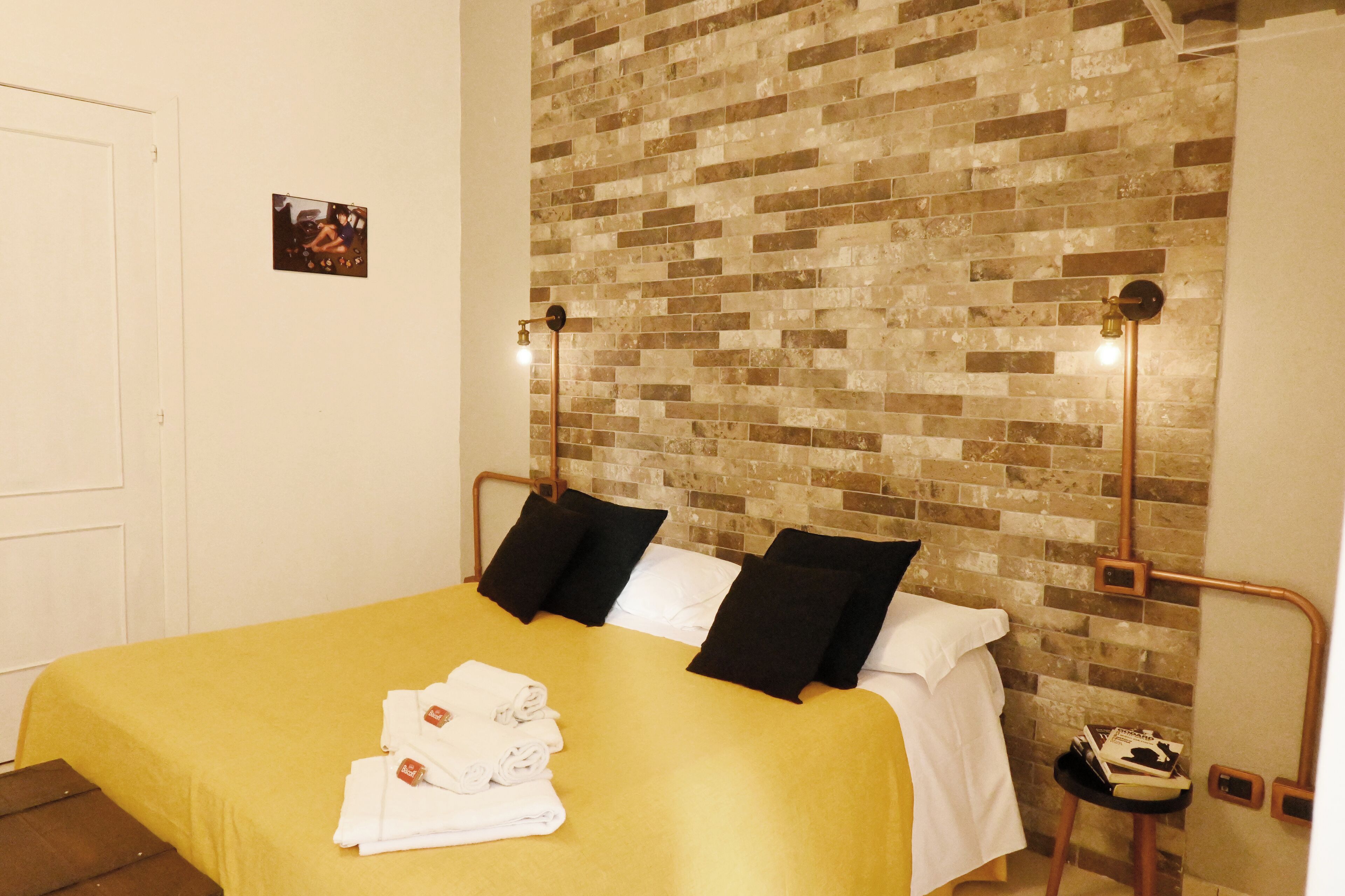 Una habitación de hotel con una pared de ladrillo, una cama con ropa de cama blanca y negra, y una mesita con un libro y un zapato.