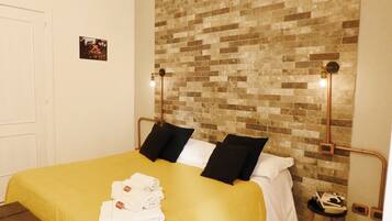 Una habitación de hotel con una pared de ladrillo, una cama con ropa de cama blanca y negra, y una mesita con un libro y un zapato.