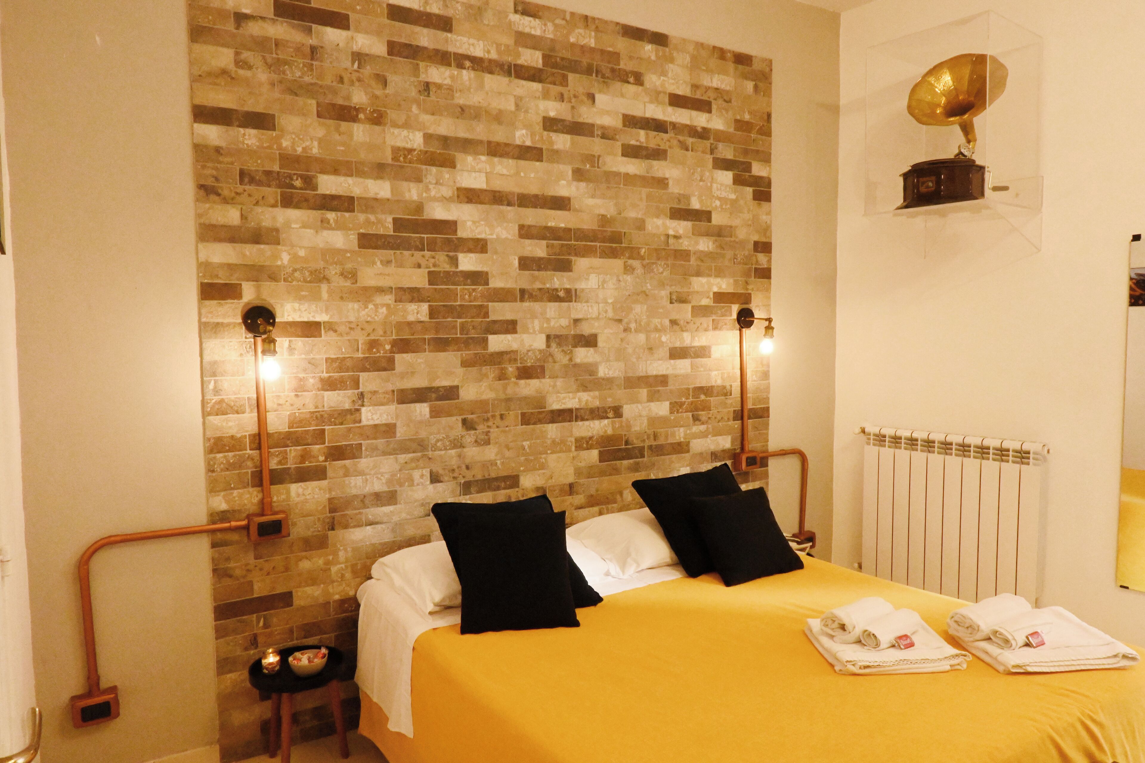 Habitación de hotel con pared de ladrillo, cama con ropa blanca, dos almohadas negras y un gramófono dorado en la pared.