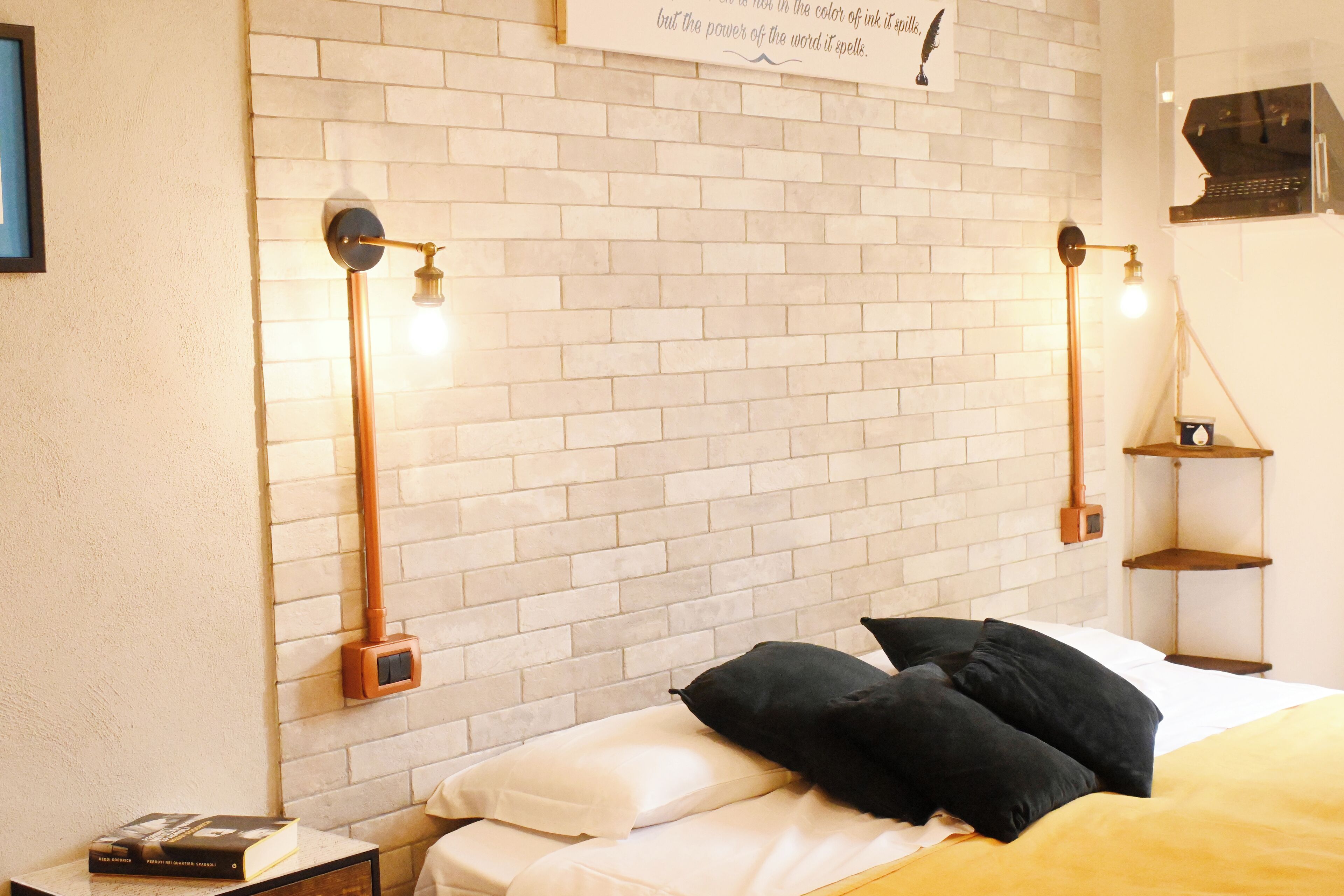 Un dormitorio ordenado con cabecera de ladrillo, una cama con ropa de cama blanco y negro y dos lámparas de lectura fijadas a la pared.