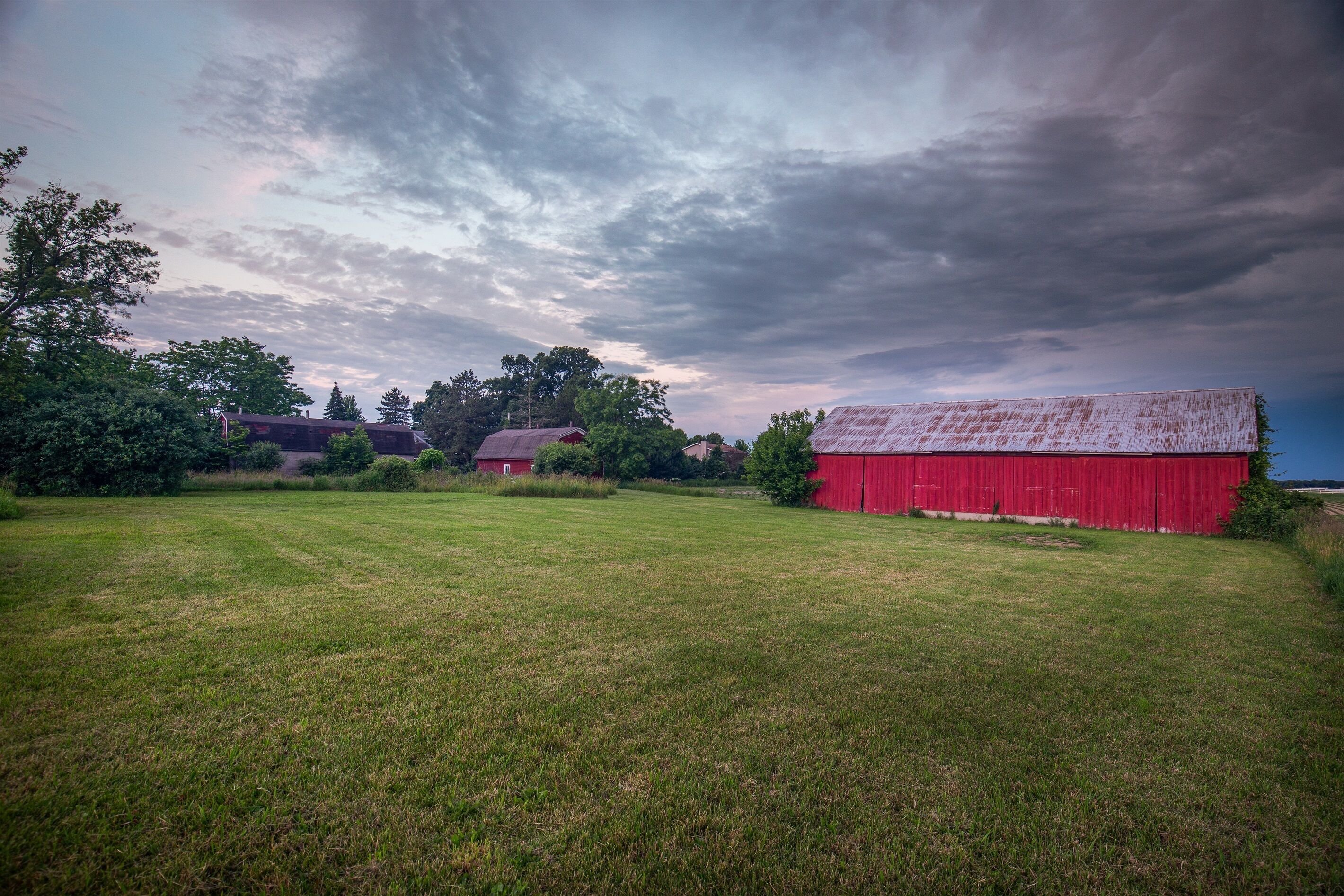 A rural landscape with a red barn, a smaller red building, and a grassy field under a cloudy sky.