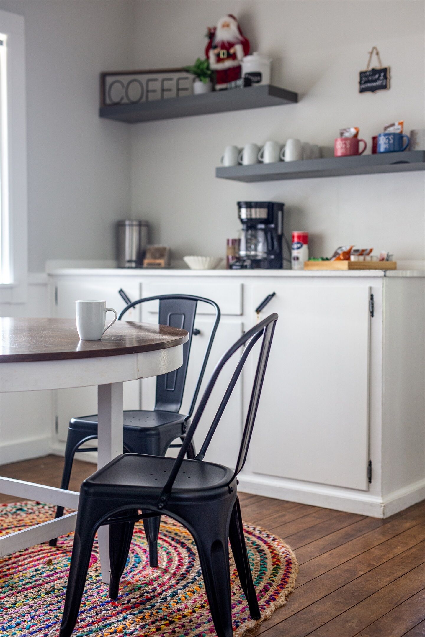 Ett kök med ett runt bord, två stolar, en hylla för kaffeprodukter och en kaffebryggare.