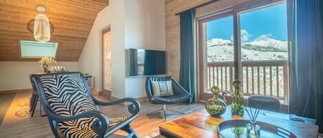 A cozy room with a wooden ceiling, a large window with a mountain view, a TV mounted on the wall, and a dining area with a table and chairs.