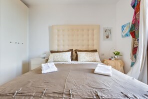 Una cama bien hecha con un cabecero color beige, almohadas blancas y una colcha marrón. Se observa una mesita de noche con una lámpara y un florero con flores.