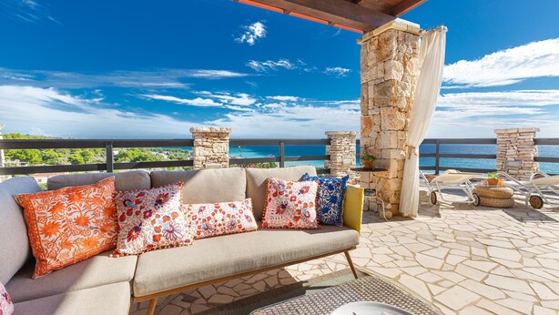 An outdoor seating area with a stone pillar, a cushioned sofa with colorful pillows, and a view of the sea.